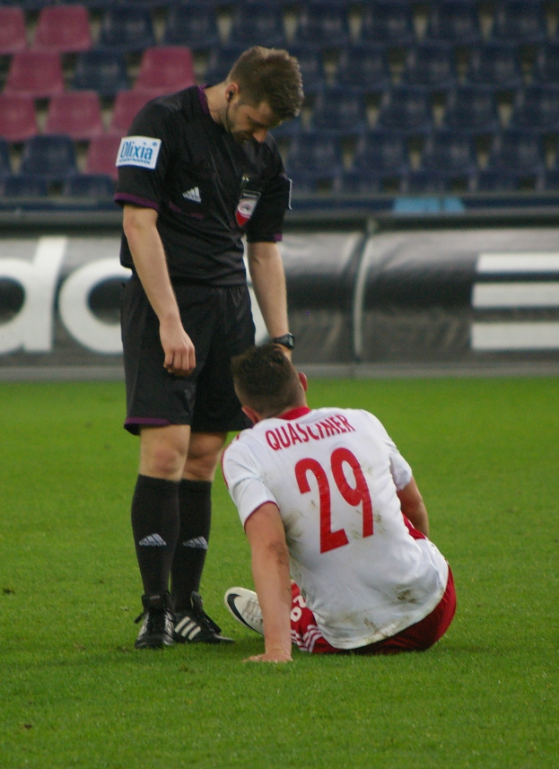 Ein Fötballspieler sitzt neben einem Schiedsrichter auf dem Boden, beide in Sportkleidung, mit Stadionplakaten und Stühlen im Hintergrund.