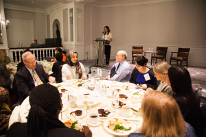 Gruppe von Menschen um einen Esstisch mit Tellern, Bestecken und Trinkgefäßen sitzen, während eine Frau in der Nähe eines Podiums mit einem Mikrofon im Hintergrund steht.