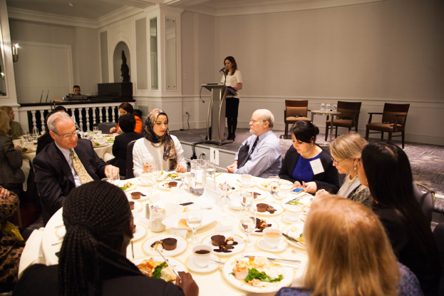 Gruppe von Menschen um einen Esstisch mit Tellern, Bestecken und Trinkgefäßen sitzen, während eine Frau in der Nähe eines Podiums mit einem Mikrofon im Hintergrund steht.