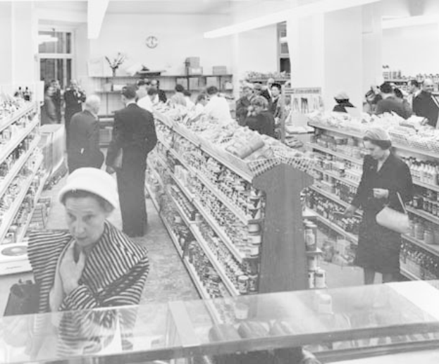 Schwarzes und weißes Foto von Menschen, die in einem Supermarkt einkaufen, mit Lebensmitteln auf Regalen, einige halten Taschen, eine Uhr an der Wand und Deckenbeleuchtung.