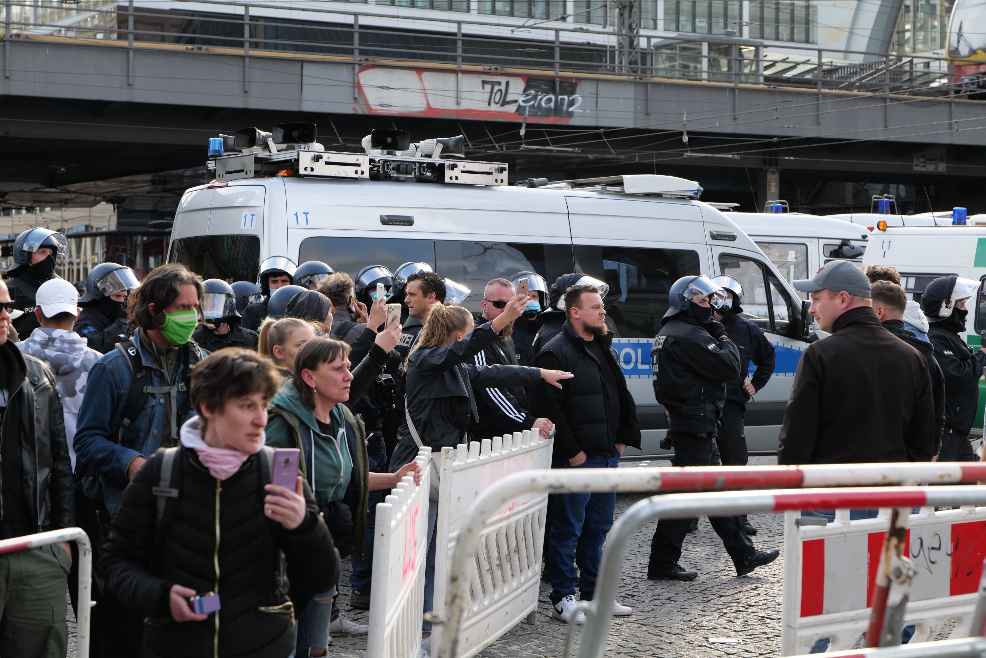 Gruppe von Menschen vor Polizeiwagen mit Absperrungen, einige tragen Helme und halten Telefone, in der Nähe einer Brücke und Gebäuden in Berlin.