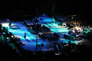 Eine Gruppe von Menschen, die auf einer Bühne mit Lautsprechern, Scheinwerfern und Mikrofonen performs, umgeben von einer großen Menge in einem dunklen Hintergrund.