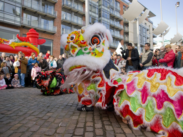 Ein lebendiges chinesisches Neujahrsfest in Amsterdam mit einem Löwentanz im Vordergrund, umgeben von einer Menge mit einigen Menschen, die Fotos machen, vor einem Hintergrund aus Gebäuden, Laternenmästen und einem klaren blauen Himmel.