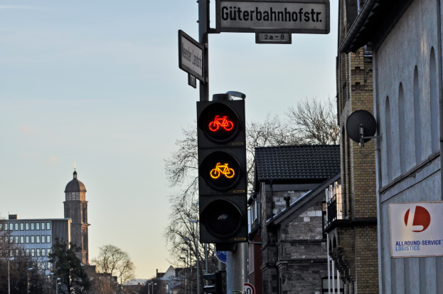 Eine Ampel mit einem Fahrradschild in einer Stadt, umgeben von Gebäuden, Bäumen, Pfählen, Plakaten und Fahrzeugen auf der Straße, mit dem Himmel im Hintergrund.