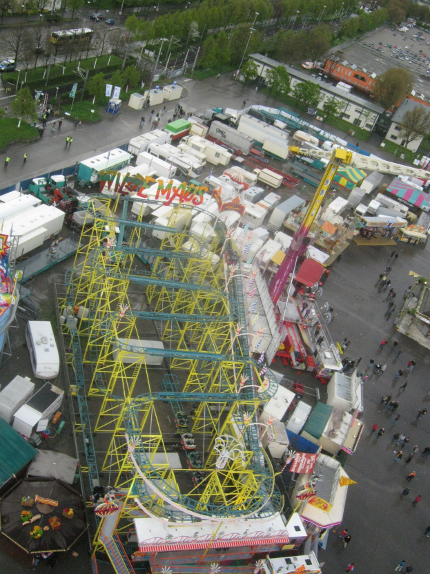 Luftaufnahme eines Vergnügungsparks mit einer Achterbahn in der Mitte, umgeben von verschiedenen Fahrgeschäften, Fahrzeugen, Menschen, Bäumen, Pfählen und Gebäuden.
