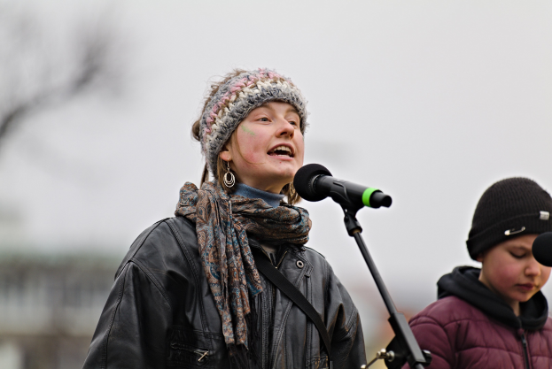 Eine Frau in schwarzer Jacke, Schal und Mütze singt in ein Mikrofon auf der Bühne, neben ihr eine Person in einer maronenfarbenen Jacke und schwarzer Mütze, während eine unscharfe Menge zusieht.