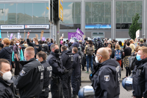 Eine Gruppe von Menschen steht vor einem Gebäude, einige halten Schilder und tragen Helme, mit einem Mast mit einem Schild im Vordergrund und einem Baum im Hintergrund, scheinbar protestierend.