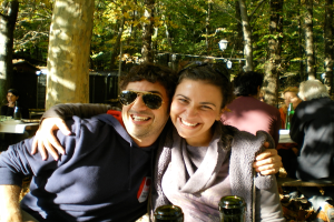 Ein Paar sitzt im Vordergrund, umarmt und lächelt, während Menschen an Tischen mit Flaschen im Hintergrund sitzen, mit Bäumen und hängenden Lämpchen im Hintergrund.