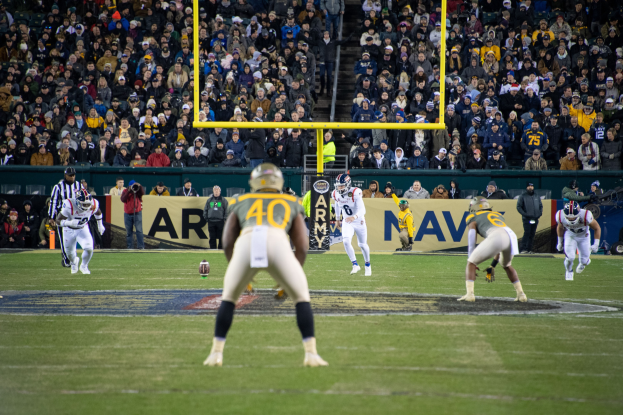 Fußballspieler in Helmen und Uniformen konkurrieren auf einem Feld mit Zuschauern auf den Rängen, während Stadionwerbung und Pfosten sichtbar sind.
