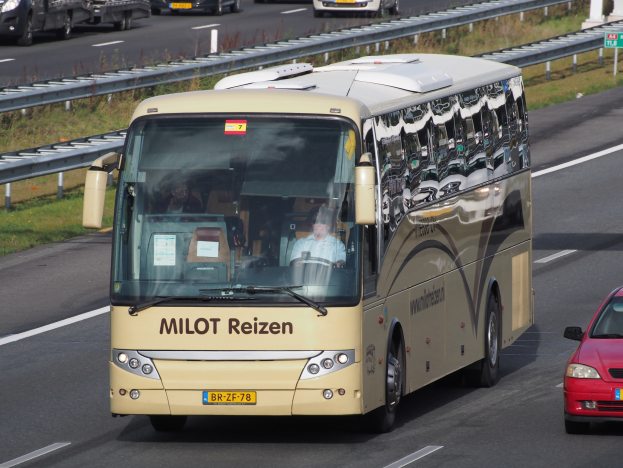 Ein Minibus mit Passagieren fährt auf einer Autobahn neben einem roten Auto, mit Geländern, Gras und texttragenden Säulen im Hintergrund.