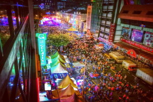 Eine volle Faschingszene mit Zelten, Geschäften, Gebäuden und Bannern, beleuchtet von Lichtern im Hintergrund.