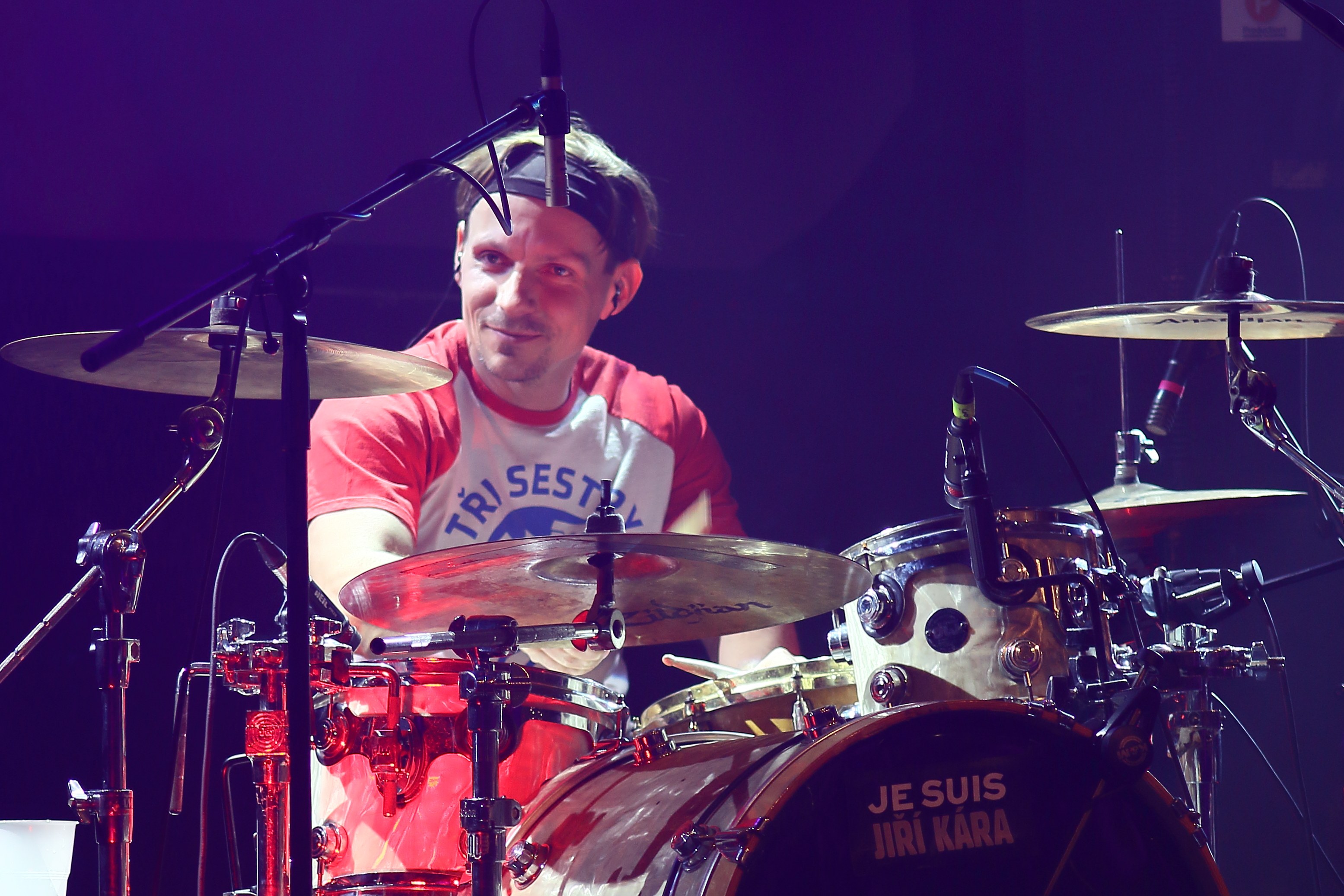 Ein Mann in einem roten und weißen T-Shirt spielt Schlagzeug auf der Bühne und lächelt während eines Konzerts mit mehreren Mikrofonen vor ihm.