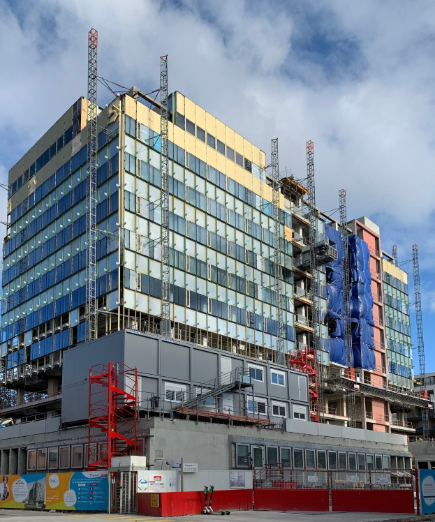 Großes Gebäude im Bau in einer städtischen Umgebung, mit Glasfenstern, Metallrahmen, einer Treppe, einem Zaun, einer Tafel, Polen, einem Straßenmast, umliegenden Bäumen und einem bewölkten Himmel.