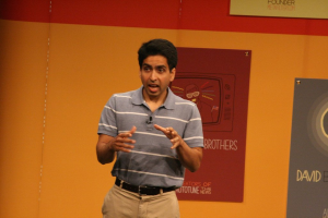 Ein Mann steht auf der Bühne und spricht mit einer Wand voller Plakate hinter ihm.