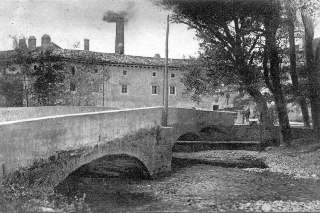 Eine Stadtansicht mit Gebäuden, Bäumen und einer Brücke im Vordergrund, Rauch aus einem Schornstein, unter einem Himmel mit Wasser am Boden.