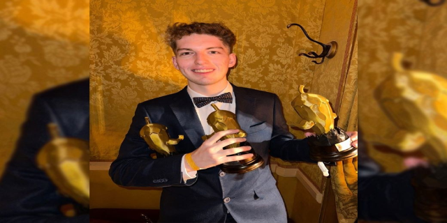 Ein Mann in einem Smoking hält eine Oscar-Statue, lächelt vor einer Wand, mit einem Wasserzeichen auf dem Bild.