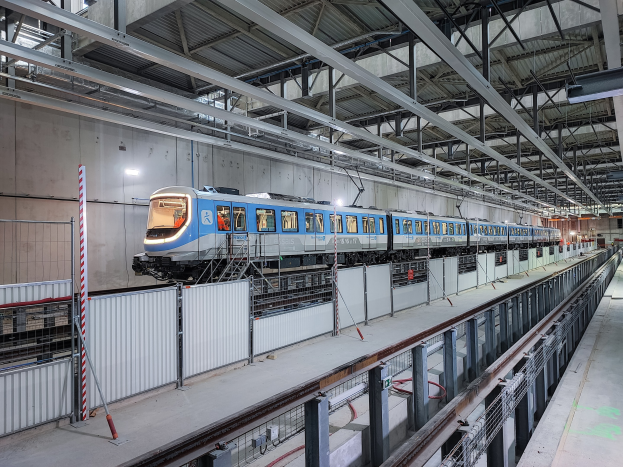 Ein blauer und weißer Zug fährt durch eine große Indoor-Station mit einer Plattform auf der rechten Seite und einer Wand im Hintergrund, unter einer Decke mit Stangen und Lichtern.
