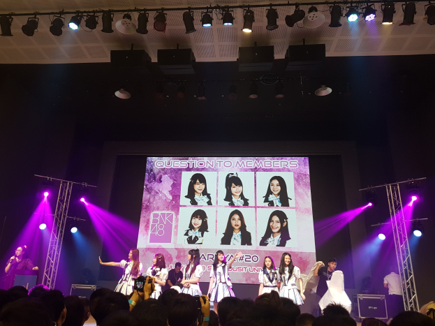 Eine K-Pop-Gruppenleistung auf der Bühne mit einem Sänger, der ein Mikrofon hält, und einem Musiker, der ein Instrument spielt, während die Menge unten aufzeichnet und ein großes Display BK48-Mitglieder anzeigt.