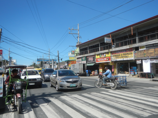 Busy street with cars, motorcycles, a zebra crossing, electric poles with wires, buildings with name boards, and a clear blue sky.