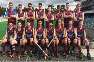 Eine Gruppe von Männern, die auf einem Feld mit Hockeystöcken feiern, Bäume und ein Stadion mit Zuschauern im Hintergrund.