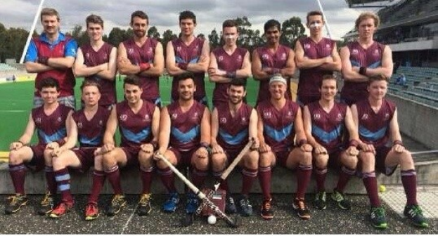 Eine Gruppe von Männern, die auf einem Feld mit Hockeystöcken feiern, Bäume und ein Stadion mit Zuschauern im Hintergrund.
