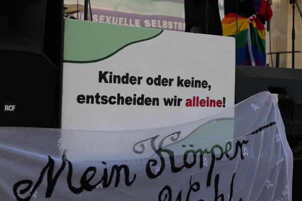 Eine Person steht auf einer Bühne mit einem Schild, auf dem "Kinder oder keine, entscheiden wir allein" steht, einer Fahne, einem Lautsprecher, einem Mikrofonständer und einem drapierten Tuch, mit einem Gebäude im Hintergrund.