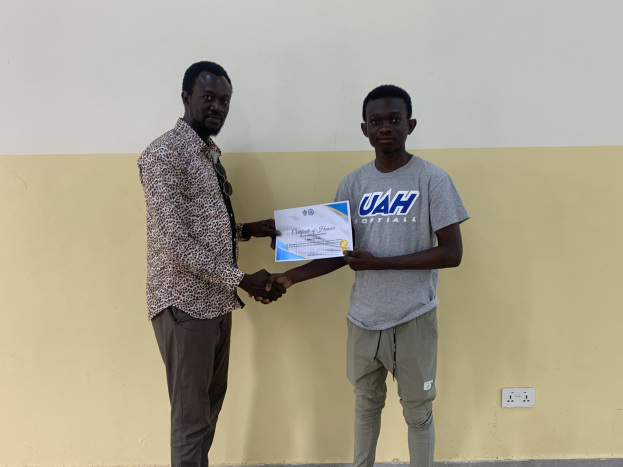 Zwei Männer schütteln sich vor einer gelben Wand die Hand, wobei der Mann links ein Zertifikat hält, das er von der Universität Ghana erhalten hat.