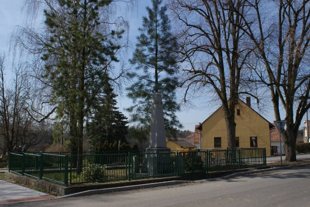 Eine Straße mit einem Holocaust-Denkmal in der Mitte, umgeben von einem Metallzaun, umgeben von Grün, Häusern, einem Straßenpfahl und einem bewölkten Himmel.