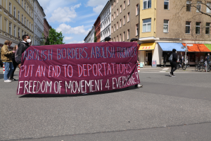 Eine Gruppe von Menschen marschiert mit einem Banner, auf dem "Abolish Borders, Abolish Frontiers, Put an End to Deportations, Freedom of Movement 4 Everyone" steht, durch eine Straße mit Gebäuden, Bäumen, Fahrrädern und einer bewölkten Landschaft im Hintergrund.