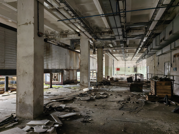 Ein verlassenes Gebäudeinnere mit Schutt, einschließlich gebrochener Holz- und Metallobjekte, die auf dem Boden, Säulen, Fenstern mit Jalousien, einer Wand im Hintergrund und einer Decke mit Rohren verstreut sind.
