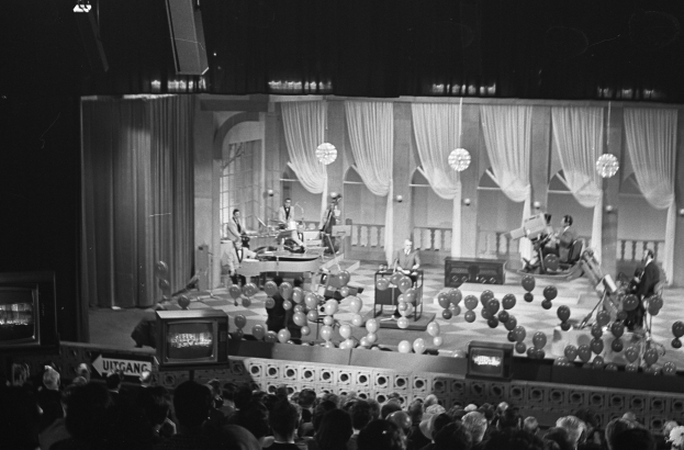 Schwarzes Bild einer Gruppe von Menschen auf einer Bühne mit Ballons und Fernsehern im Hintergrund.