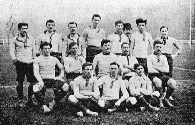 Ein Schwarz-Weiß-Foto einer Fußballmannschaft aus den 1920er Jahren, bei dem einige Spieler stehen und andere auf dem Boden sitzen, vor Pfählen, Bäumen und einem klaren Himmel.