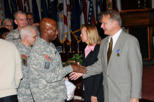 Eine Gruppe von Menschen, die mit zwei Personen Handschlag gibt, Flaggen im Hintergrund, ein Podium mit Mikrofon und eine Topfpflanze in der Nähe.