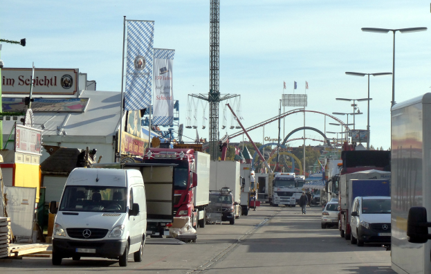 Vollgestellte Straße mit Lastwagen, Autos, Fußgängern, Laternenmasten, Bannern, einem Turm, einem Vergnügungsgerät, Bäumen und einem bewölkten Himmel.