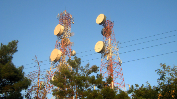 Zwei Mobilfunkmasten mit Antennen, umgeben von Bäumen und Drähten, vor einem klaren blauen Himmel.