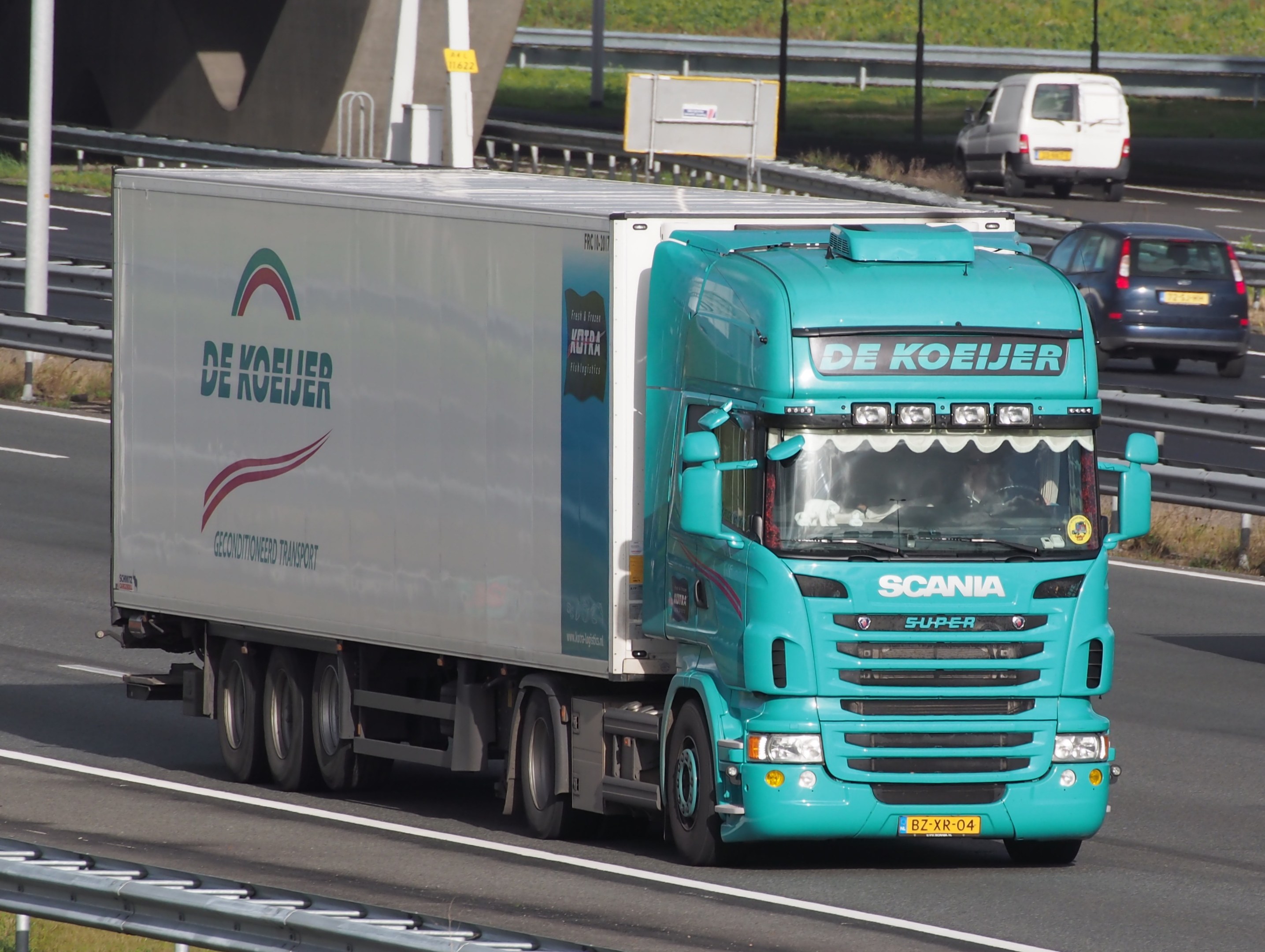 Blauer Sattelschlepper auf einer Autobahn neben einer Brücke mit Geländern, Pfählen, Brettern und Gras, mit anderen Fahrzeugen auf der Straße zu sehen.