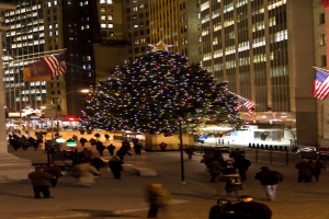 Ein Weihnachtsbaum mit Menschen in der Mitte, links ein Fahnenmast, rechts ein weiterer Fahnenmast und beleuchtete Geb√§ude im Hintergrund.