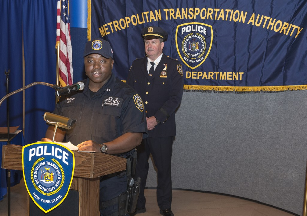 Zwei Polizisten an einem Rednerpult mit einer Fahne, einem Banner und einer Tafel mit Symbolen und Text.