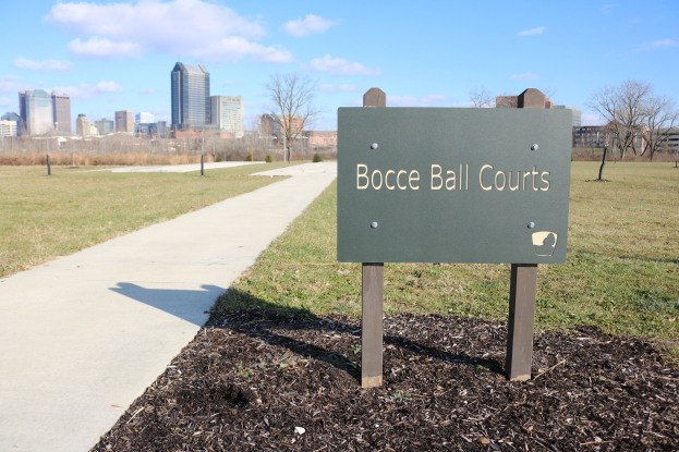 Ein Schild am Boden trägt die Aufschrift "Boccia-Plätze in Indianapolis, Indiana" in einer parkähnlichen Umgebung mit Bäumen, einem Zaun und Gebäuden im Hintergrund.