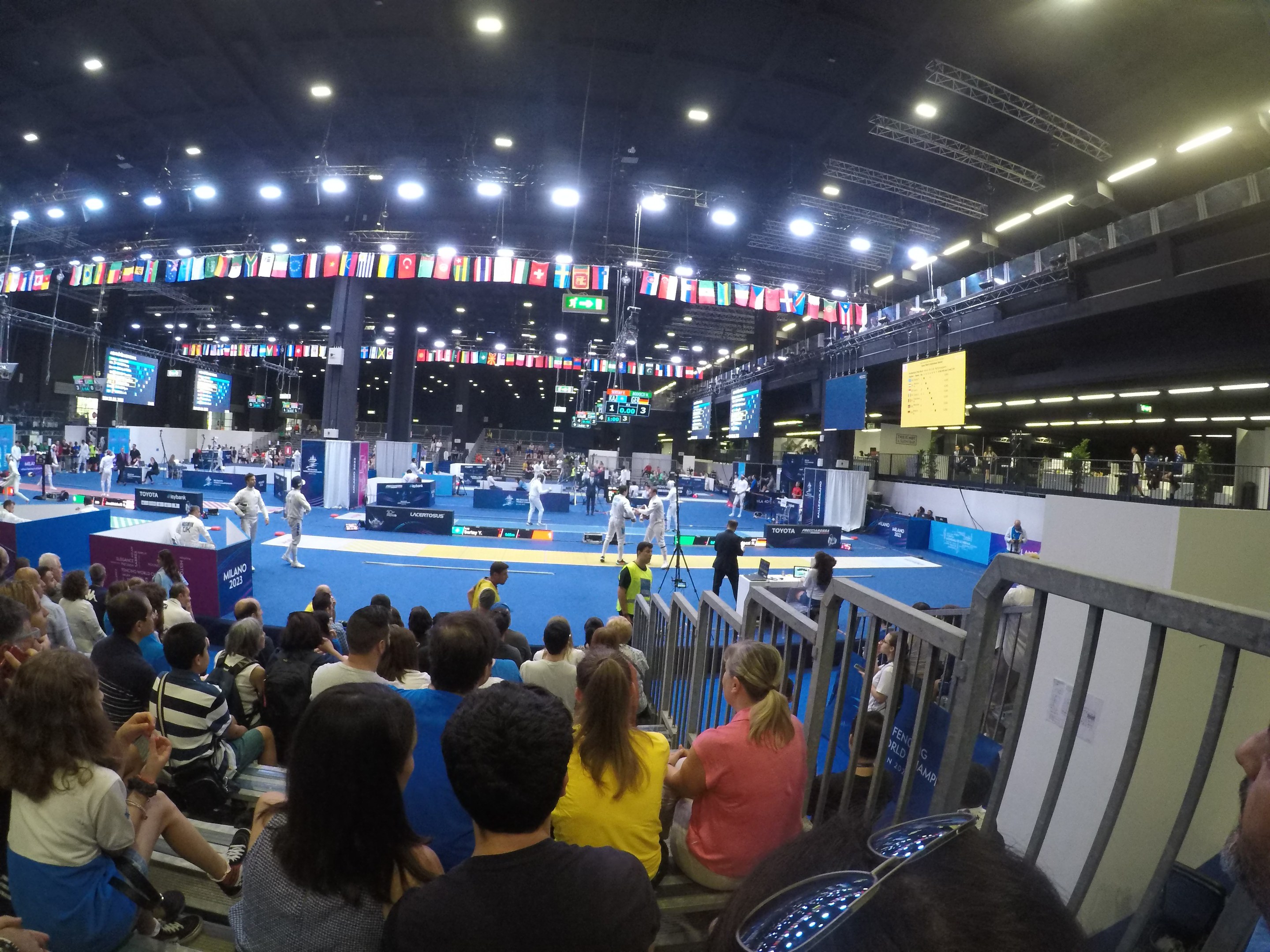 Große Menschenmenge sitzend und stehend in einer Indoor-Sportarena mit Bildschirmen, Flaggen und Deckenbeleuchtung.