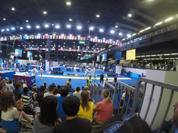Große Menschenmenge sitzend und stehend in einer Indoor-Sportarena mit Bildschirmen, Flaggen und Deckenbeleuchtung.