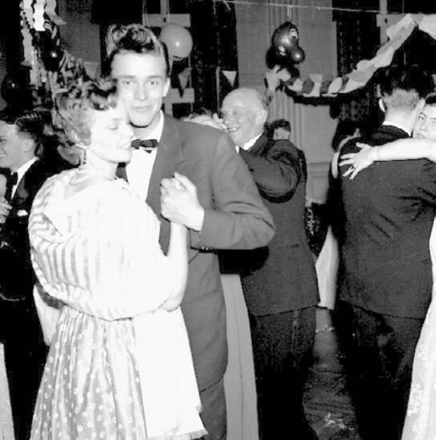 Ein Schwarz-Weiß-Foto von einem Mann und einer Frau, die auf einer Party tanzen, umgeben von lachenden Menschen und Ballons sowie Dekorationen im Hintergrund.