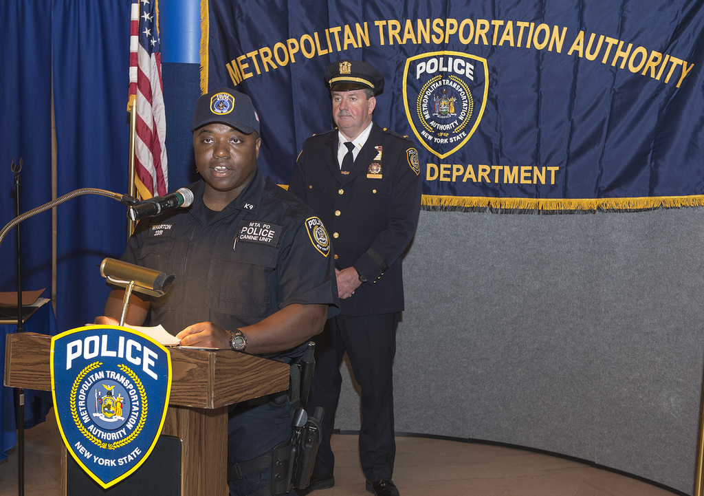 Zwei Polizeibeamte stehen vor einem Rednerpult, einer hölt Papiere und spricht in ein Mikrofon, mit einer Fahne, einem Tisch und einer Tafel mit Symbolen und Text im Hintergrund.