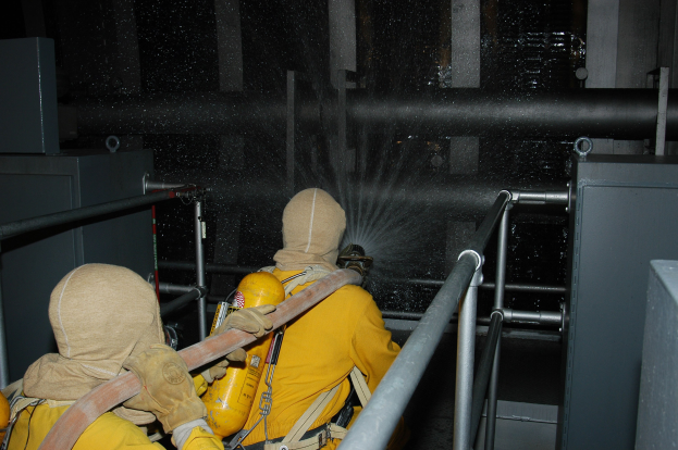 Zwei Feuerwehrleute in gelben Uniformen, Helmen und Handschuhen sprühen Wasser aus einem Schlauch auf ein Metallgeländer mit Metallstangen und anderen Gegenständen im Hintergrund.