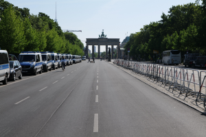 Eine lange Reihe von Polizeiwagen, die auf der Seite einer Straße vor dem Brandenburger Tor in Berlin, Deutschland, geparkt sind, mit Menschen, die Fahrräder fahren und auf der Straße stehen, sowie Barrieren und Bäume, die die Seiten säumen, und ein Bogen mit Statuen im Hintergrund.