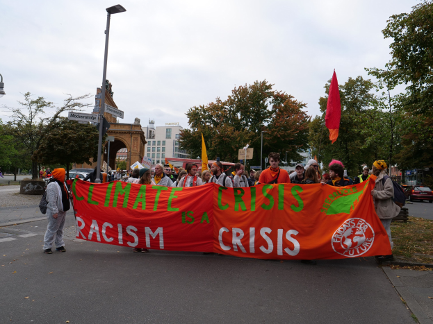 Eine Gruppe von Menschen marschiert eine baumbestandene Straße entlang und hält ein Banner mit der Aufschrift "Klimakrise ist eine Krise". Im Hintergrund sind Fahrzeuge und Gebäude zu sehen.