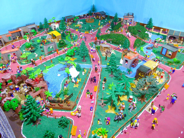 Ein Miniatur-Zoo-Modell mit Besuchern, umgeben von Bäumen, Häusern, Pflanzen, einem Zaun und einer Hintergrundwand, mit Text unten.