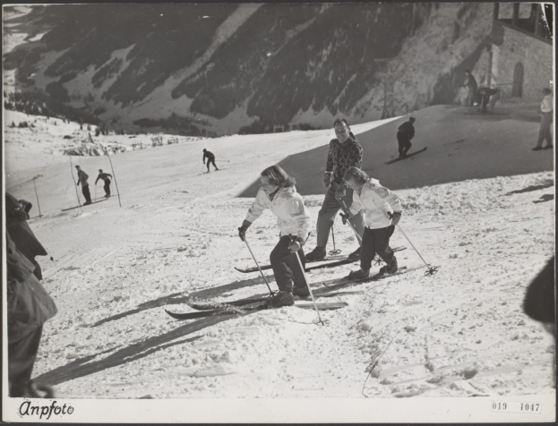 Gruppe von Menschen, die eine schneebedeckte Piste mit Skistöcken hinunterfahren, mit Hügeln und einem Gebäude im Hintergrund und Text am unteren Rand.