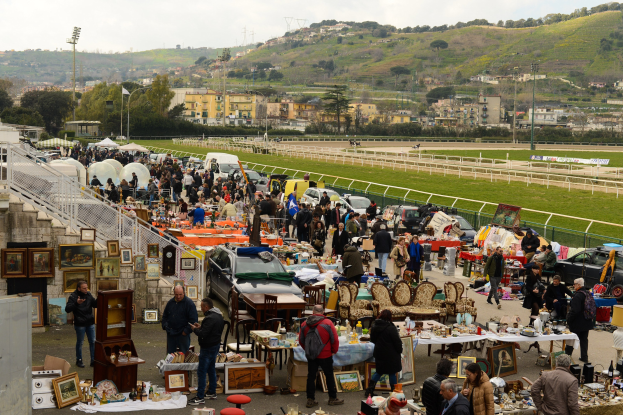 Große Gruppe von Menschen auf einem Flohmarkt mit Tischen, auf denen Gegenstände wie Rahmen und Stühle ausgebreitet sind, Fahrzeuge in der Nähe geparkt, Geländer, Treppen, Bäume, Gebäude, Laternenpfähle, Hügel und ein bewölkter Himmel im Hintergrund.