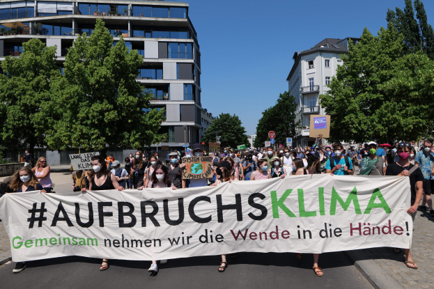 Gruppe von maskierten Menschen mit einem Banner "Aufbruchsklima" vor einem Gebäude mit Bäumen und klarem Himmel.
